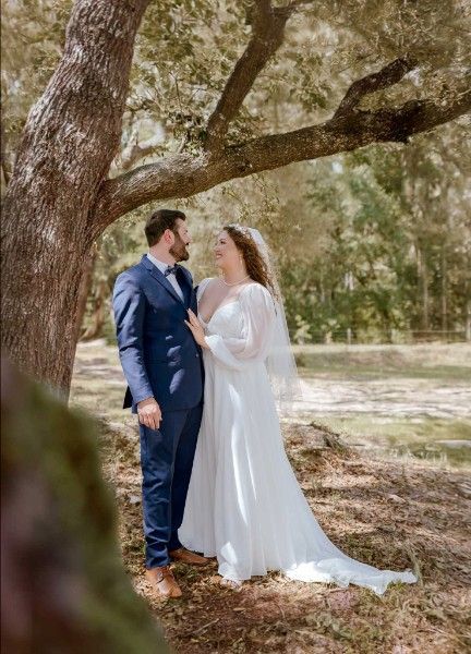 A romantic wedding moment captured under a large oak tree, with the couple in classic white and navy attire.