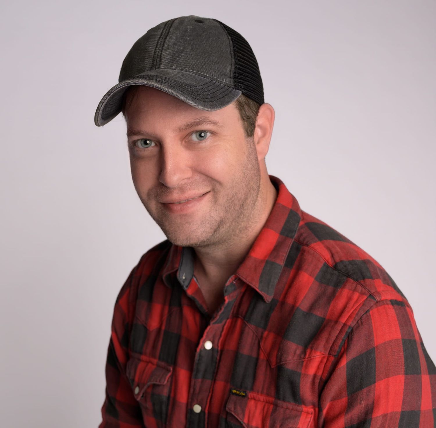 Casual headshot of man wearing red plaid shirt and baseball cap against gray background.