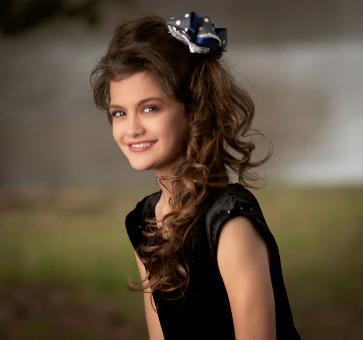 Portrait of person wearing black dress with curled hair and decorative hair accessory.
