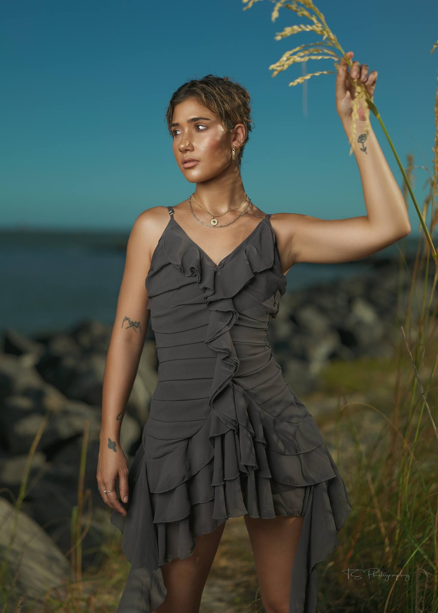 Coastal fashion portrait in grey ruffled dress with wheat stalks against blue sky.