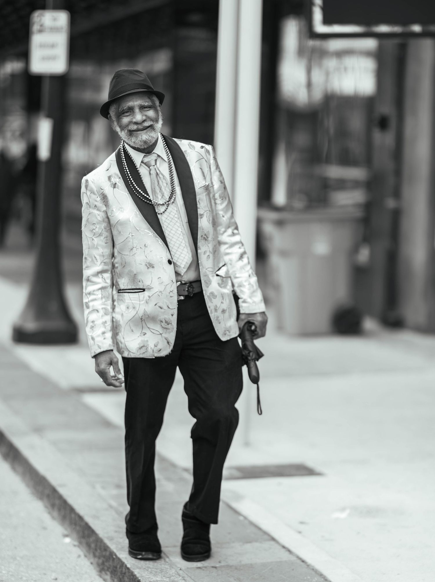 Elderly person in a stylish patterned white blazer, dark hat and pants walks along a city sidewalk in black and white.