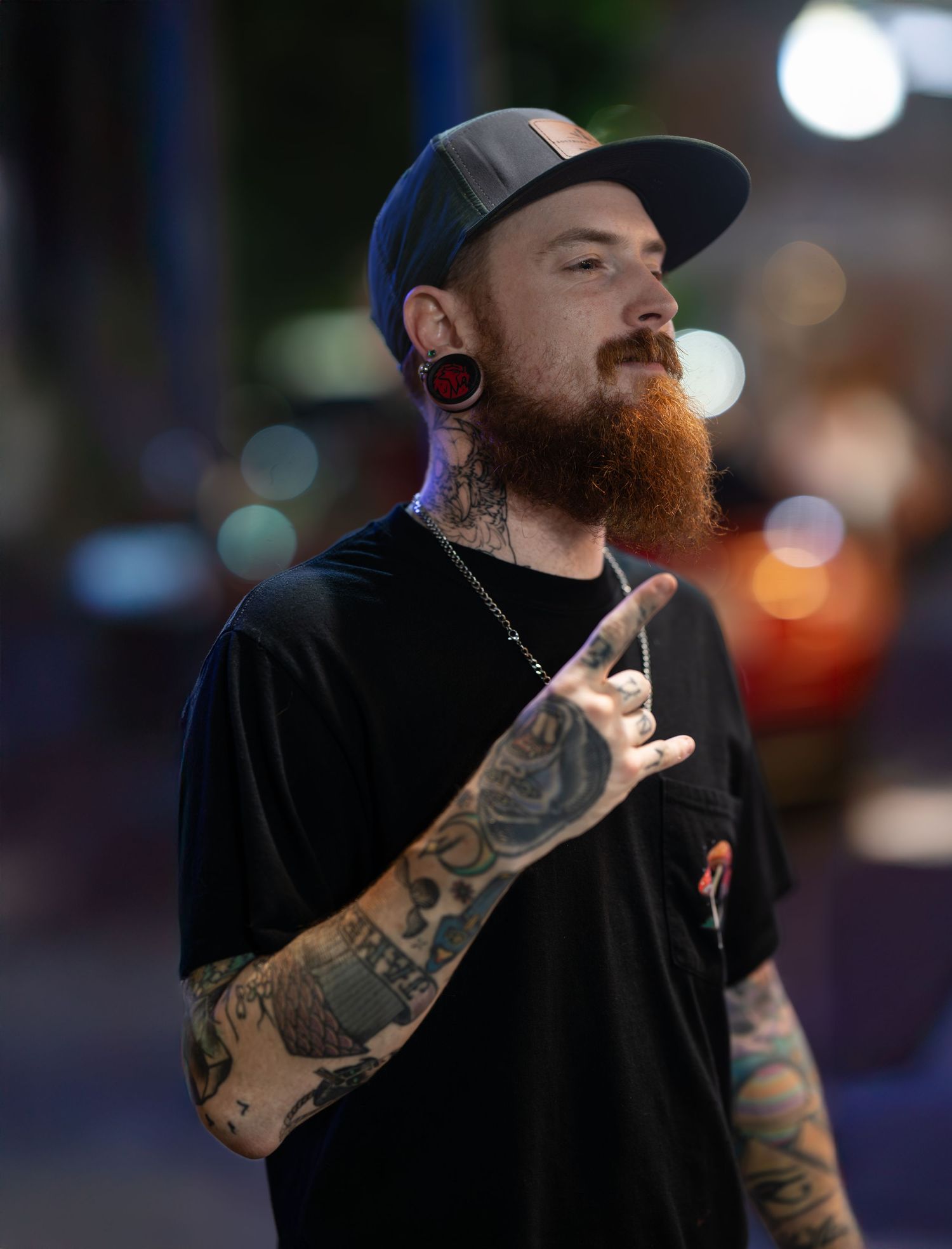 A person with a full beard, ear gauges and tattoos wearing a baseball cap and black t-shirt gestures while standing outdoors.
