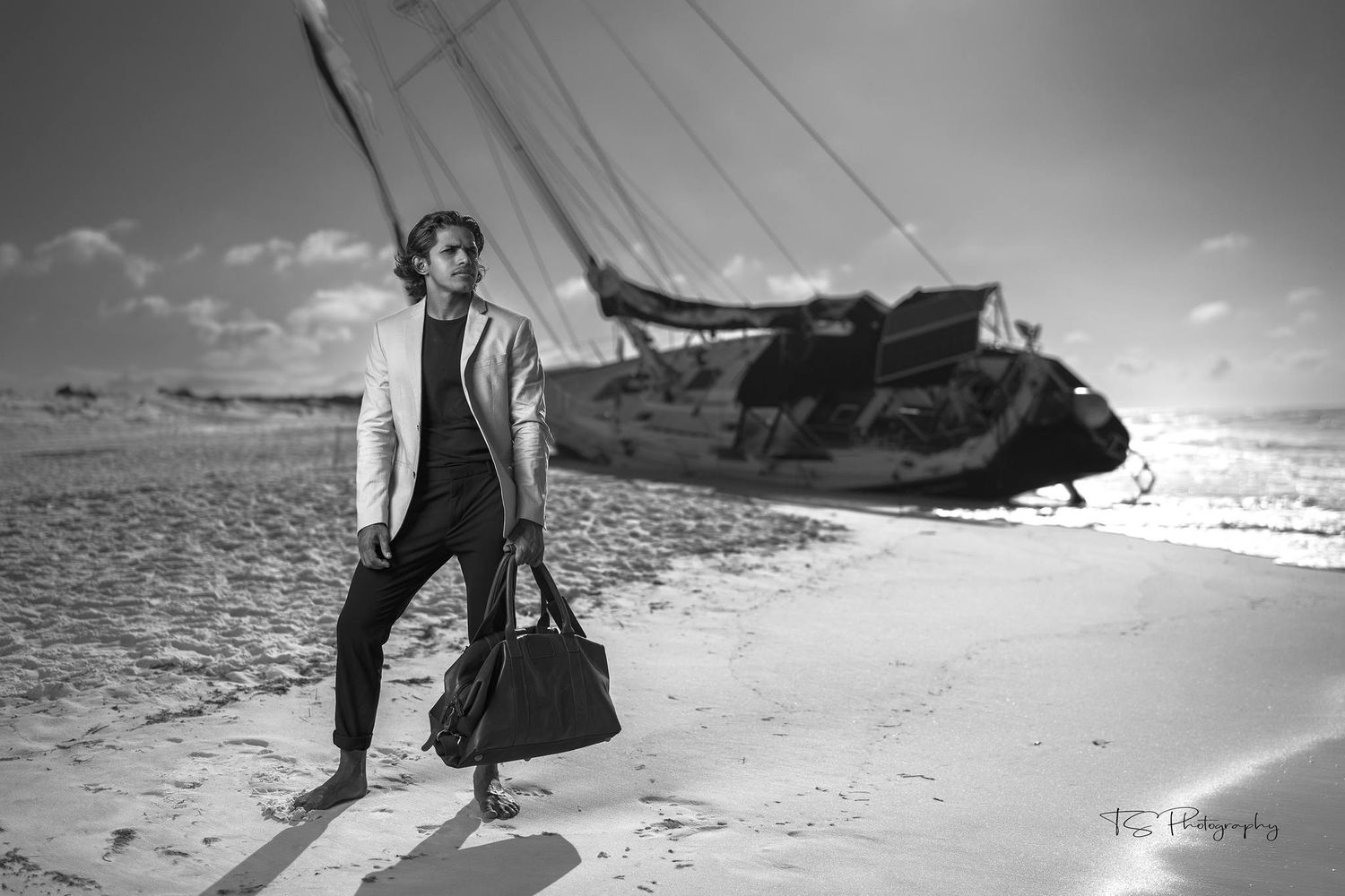 Artistic black and white photo of person with luggage standing near shipwreck on beach.