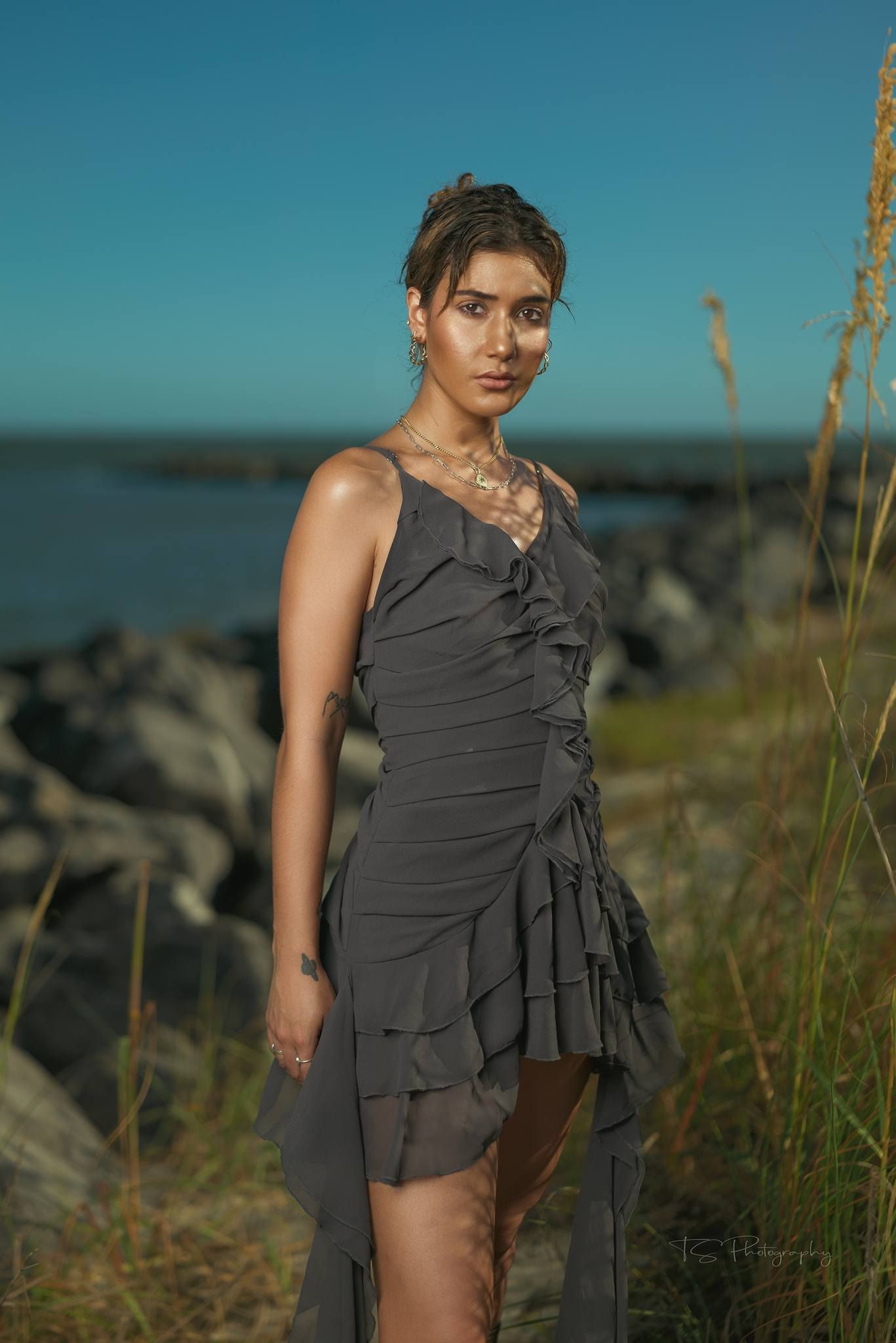 Seaside fashion shoot featuring grey ruffled dress against rocky coastal background.