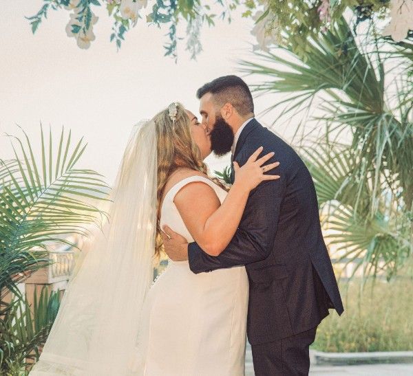 A couple shares a romantic kiss surrounded by tropical palm fronds and greenery on their wedding day.