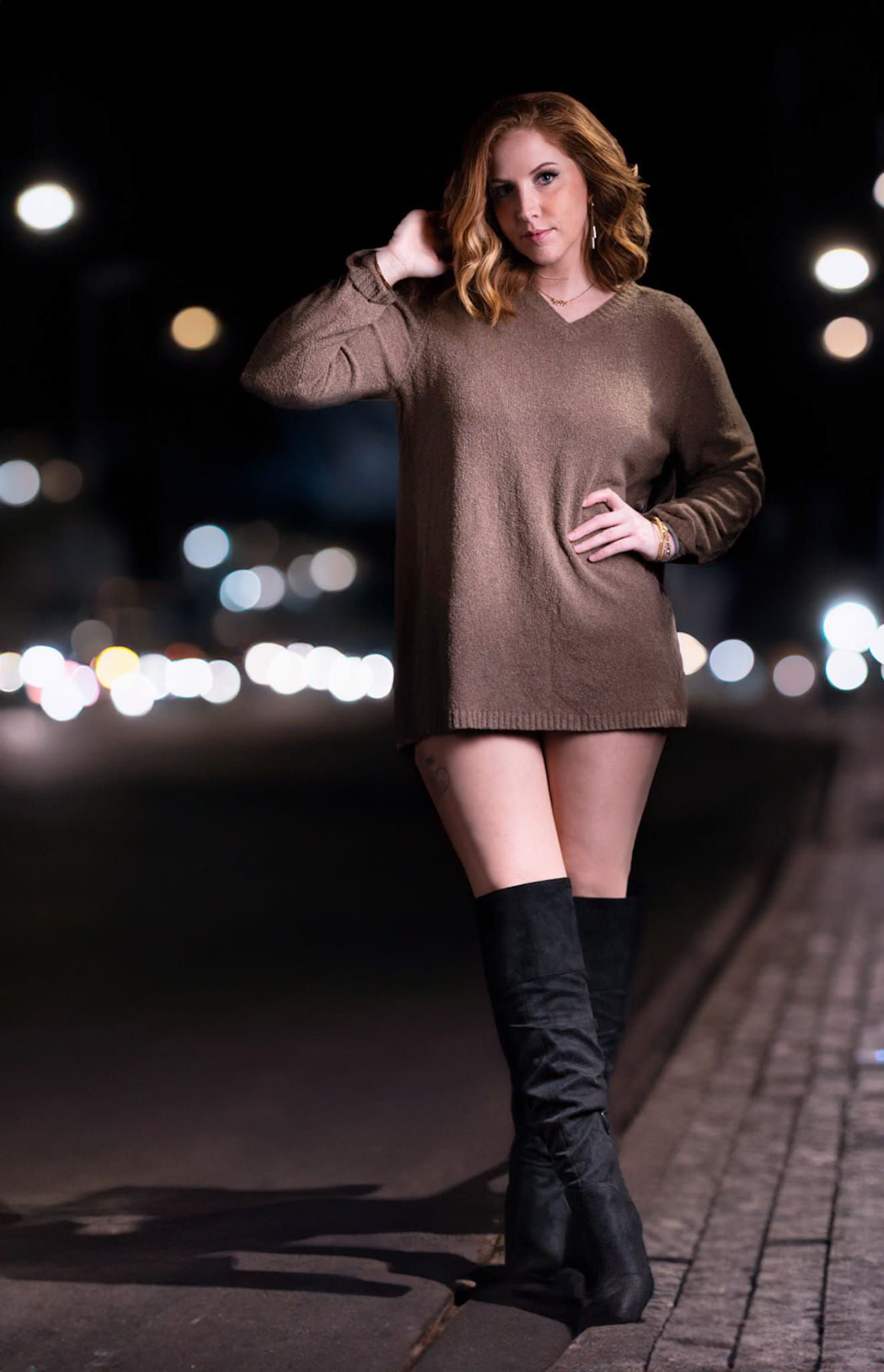 A figure in an oversized brown sweater and tall boots poses at night with bokeh lights in the background.