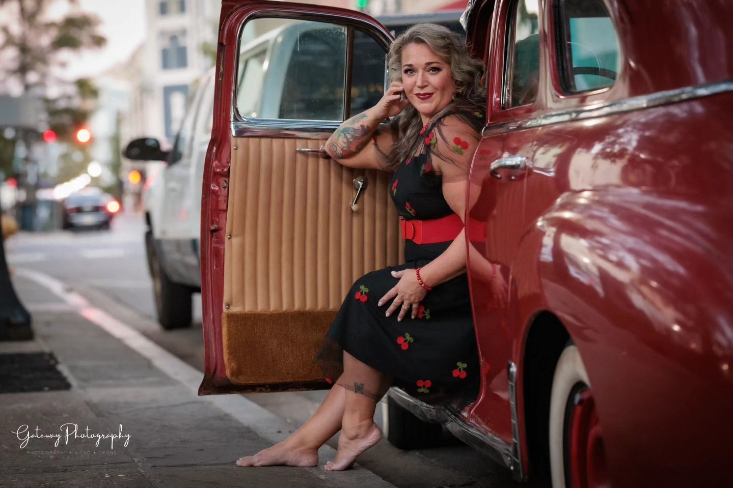 Model poses in vintage red car wearing black dress and bare feet.