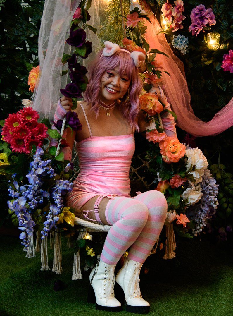Cosplay scene with pink-haired person in pink dress surrounded by colorful flowers.