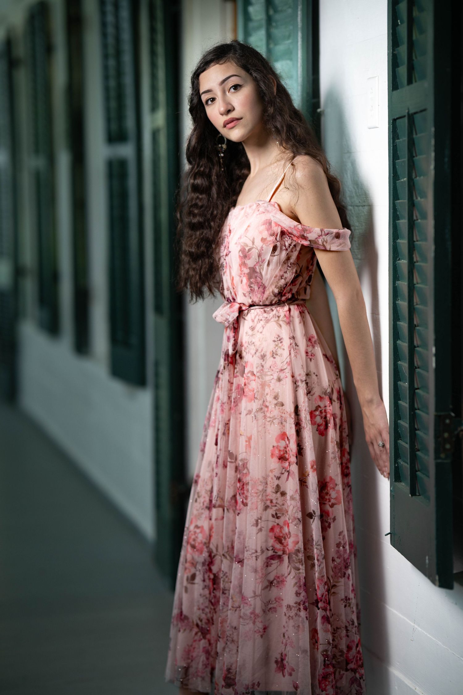 Young woman in a floral pink off-shoulder maxi dress poses gracefully in a corridor with green shutters.