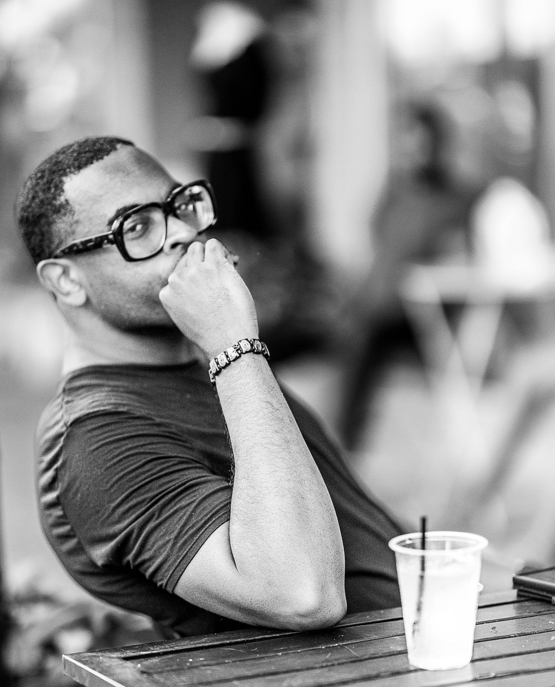 Black and white portrait of person in glasses resting hand on chin at outdoor table.