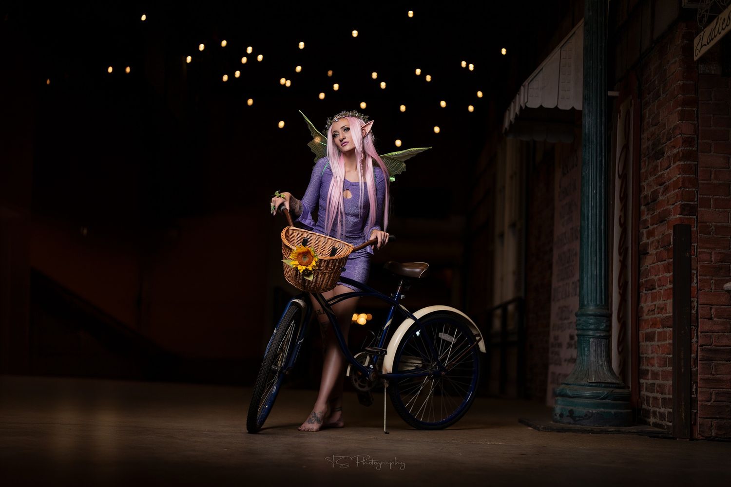 Purple-haired biker posing with motorcycle under twinkling lights at night.