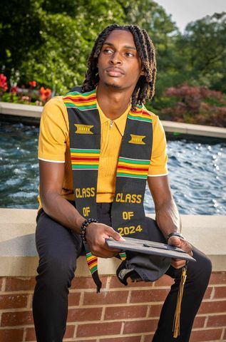 Graduate in yellow and green kente stole sits by pool holding diploma for class of 2023.