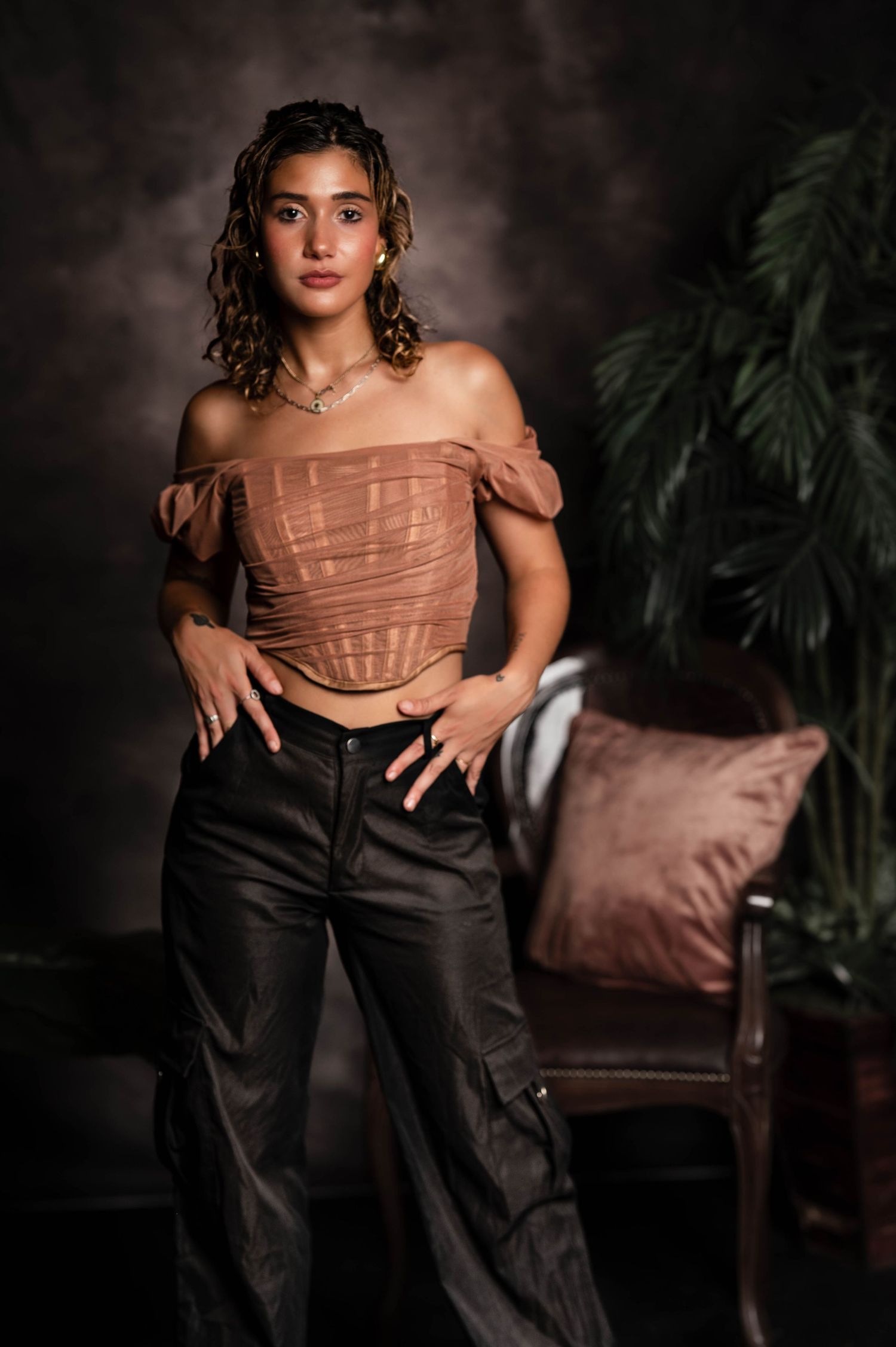 Stylish portrait with off-shoulder corset top and dark pants against moody backdrop with indoor plants.