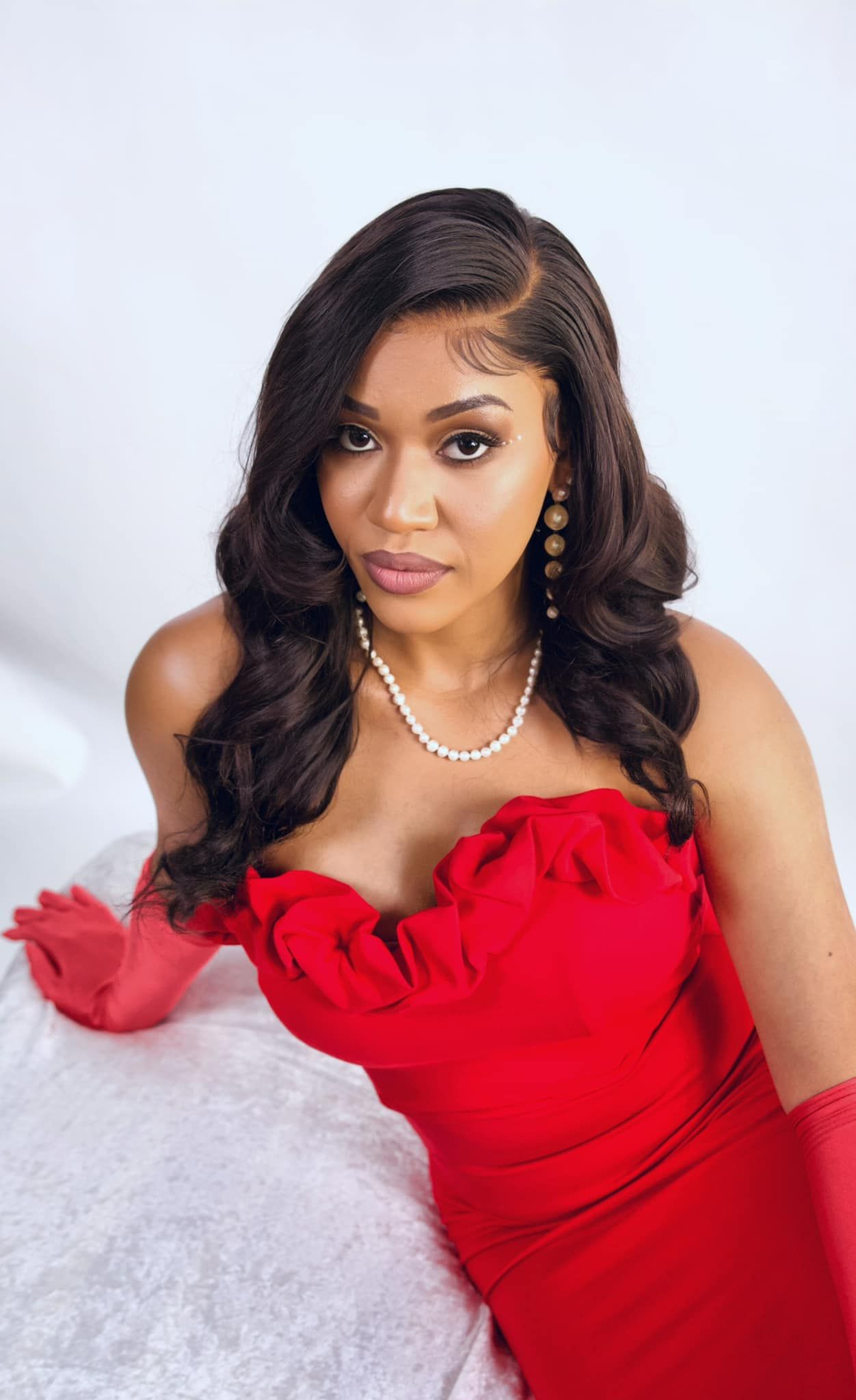 A glamorous portrait in a red strapless gown with pearl necklace and matching earrings against a white backdrop.