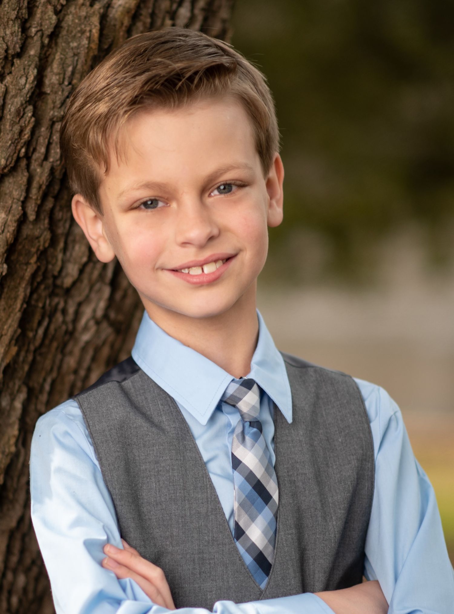 Young person in dress shirt, tie and grey vest poses by tree trunk.