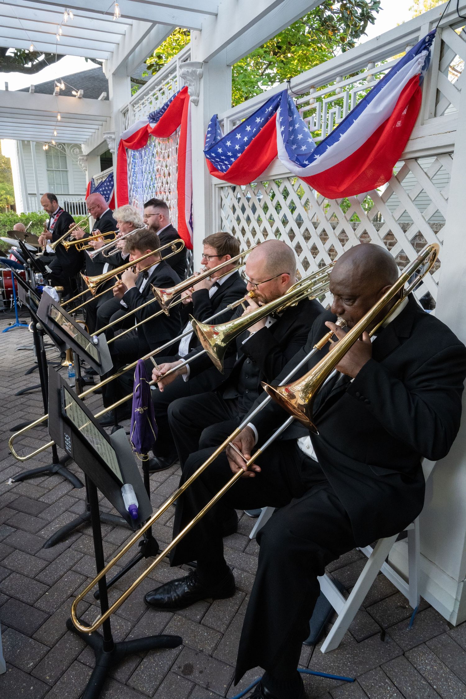 Trombone players in black suits perform outdoors under patriotic red, white, and blue bunting decorations.