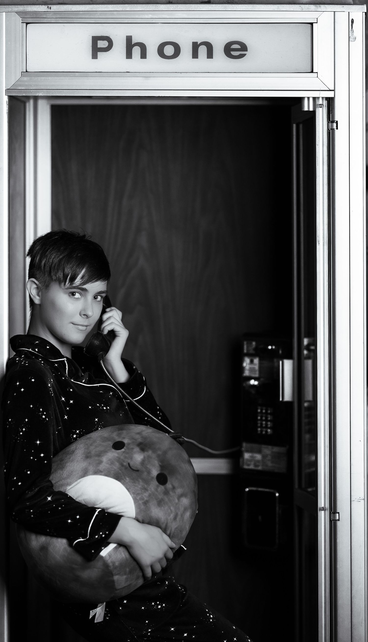 A black and white photograph shows a person in a phone booth wearing a spotted sweater and holding a drum.