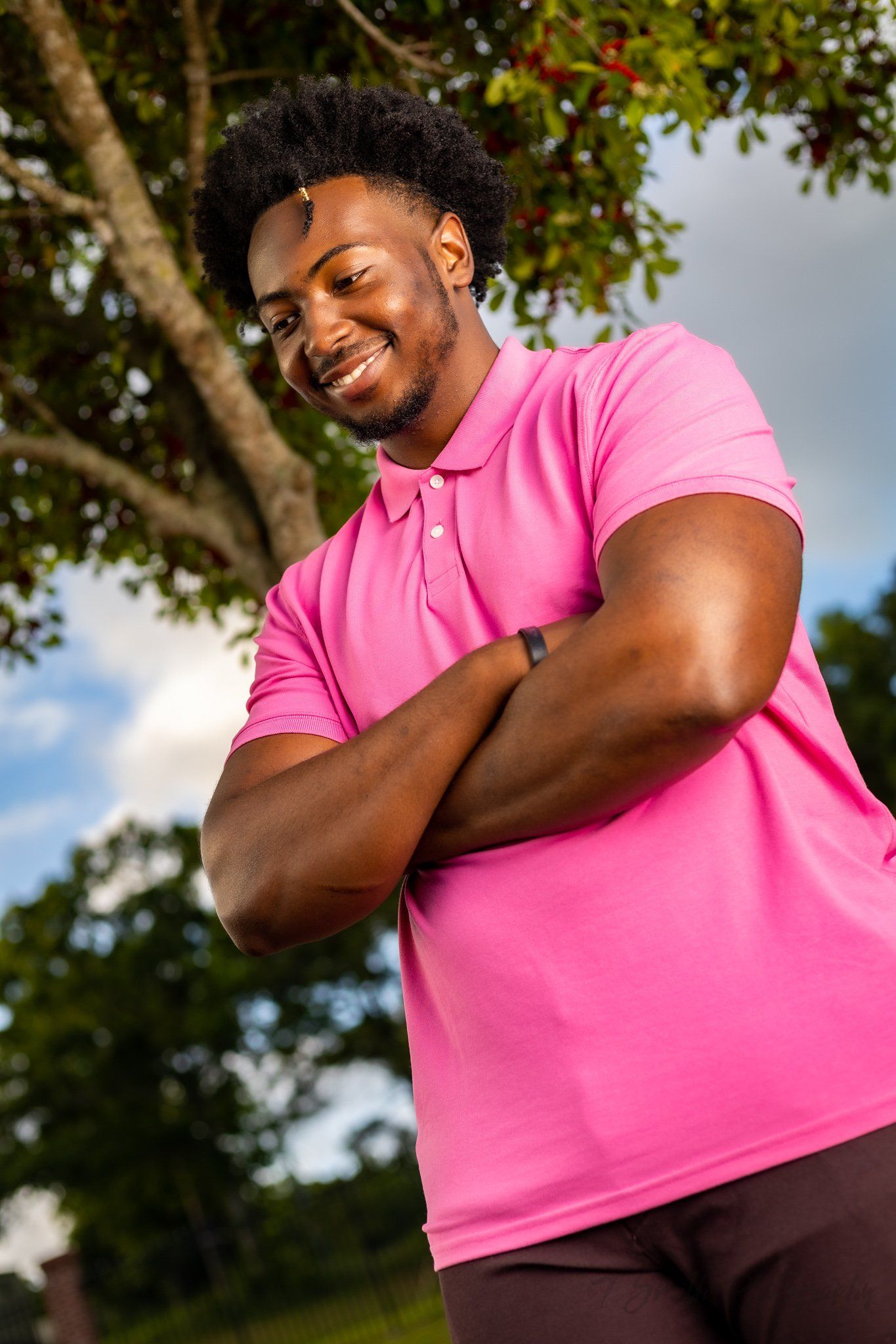 Outdoor portrait in a bright pink polo shirt against natural greenery and blue sky.