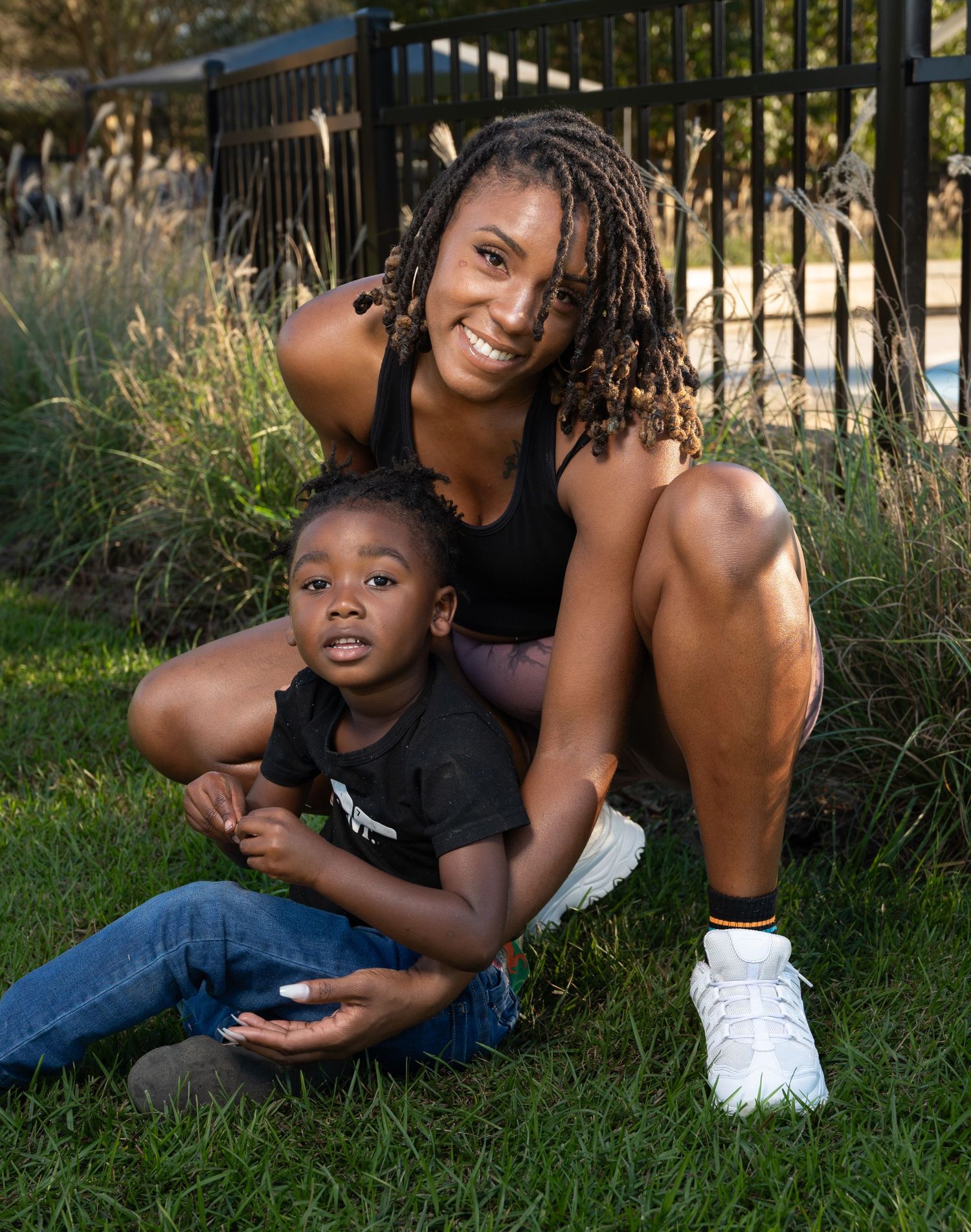 A happy parent and child share a tender moment while sitting on grass near a fence in a park.