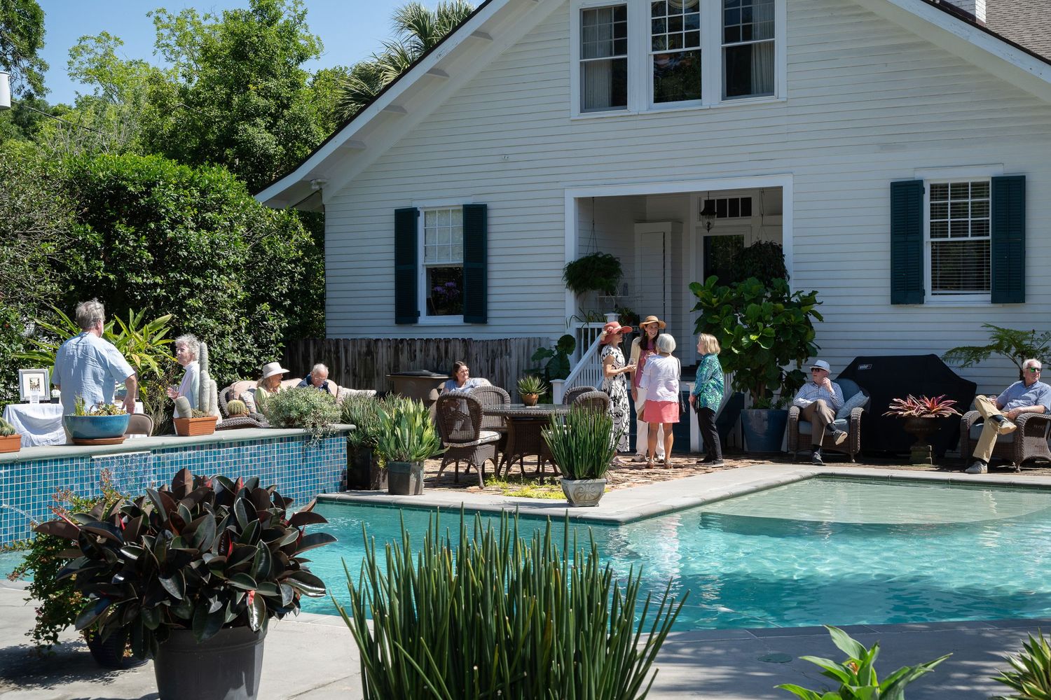 Guests gather poolside at a white house with green shutters during an outdoor summer party.