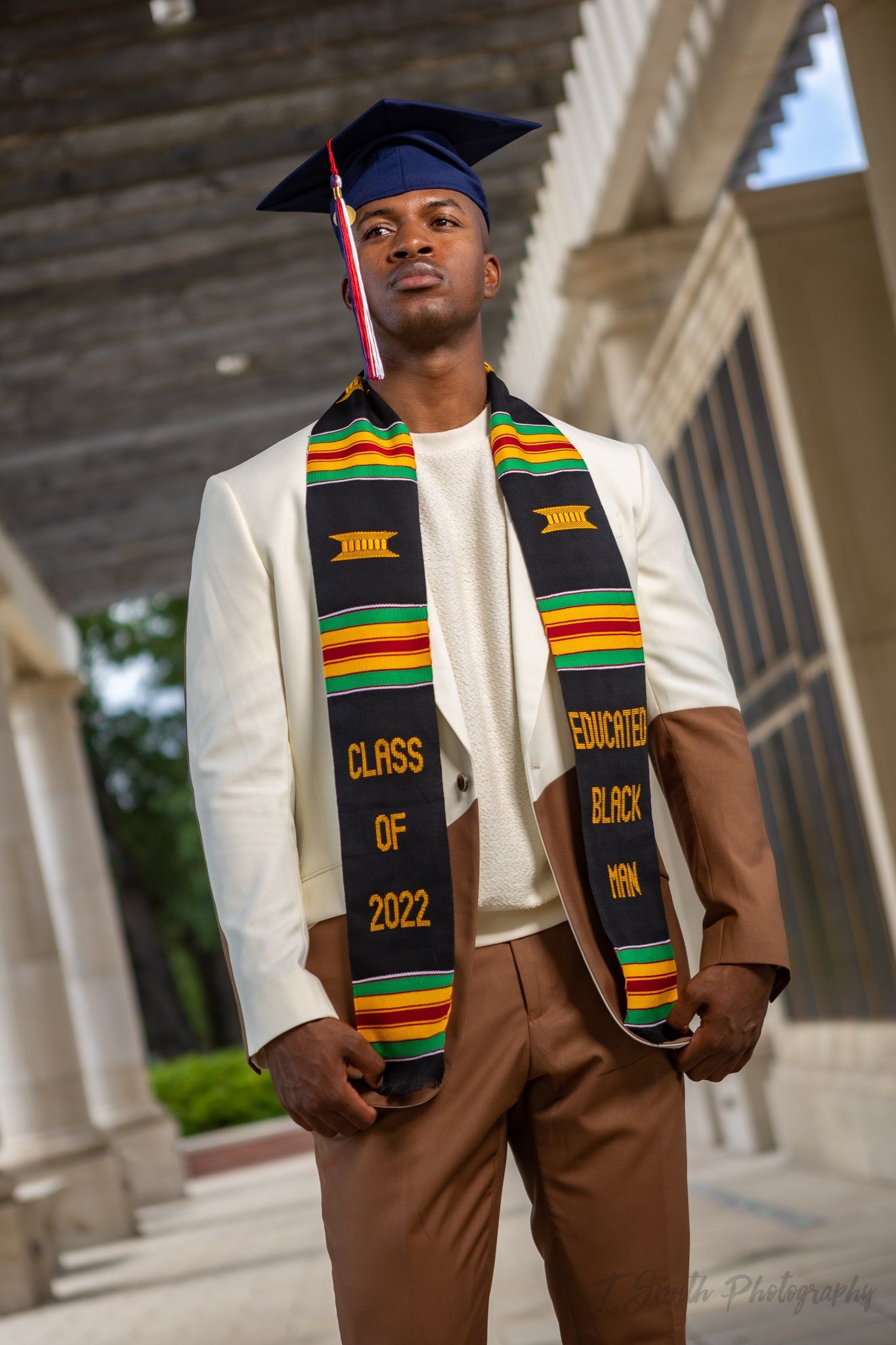 Graduate in white blazer wearing traditional kente stole and cap poses near classical columns.