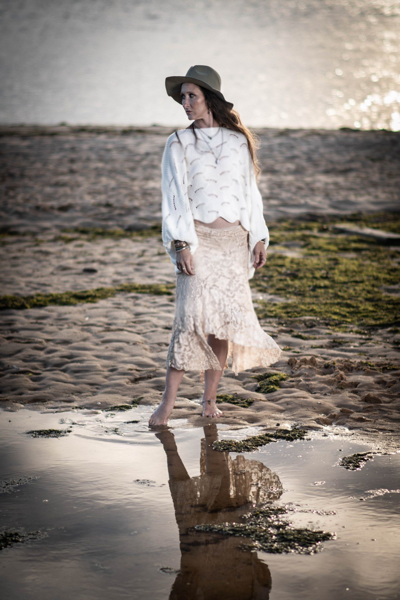 Romantic vintage-style portrait on beach at sunset with lace skirt, white blouse and floppy hat creating reflection in tide pool.