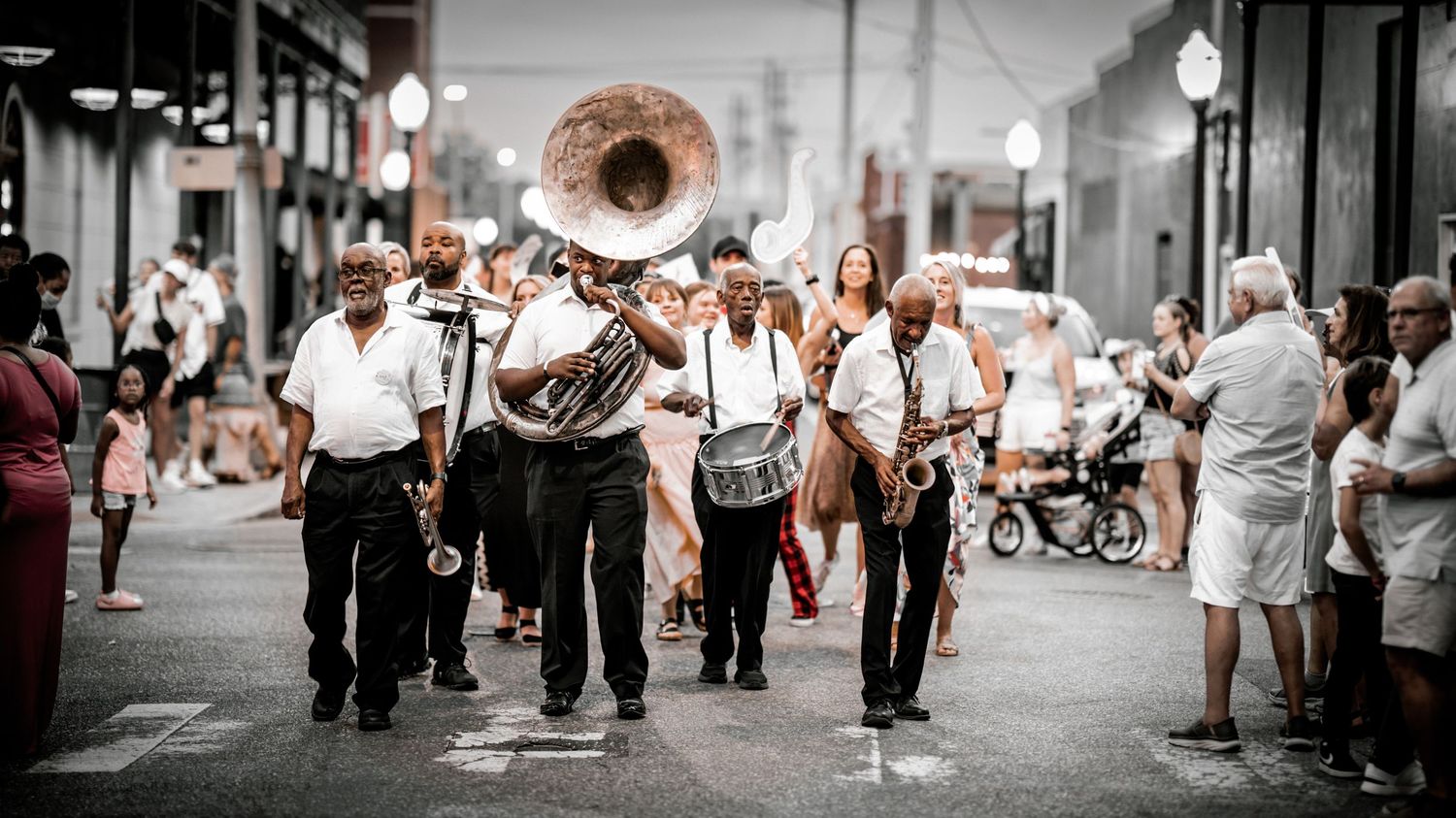 A brass band parade marches down a New Orleans street, led by a tuba player, with onlookers watching from the sidewalk.