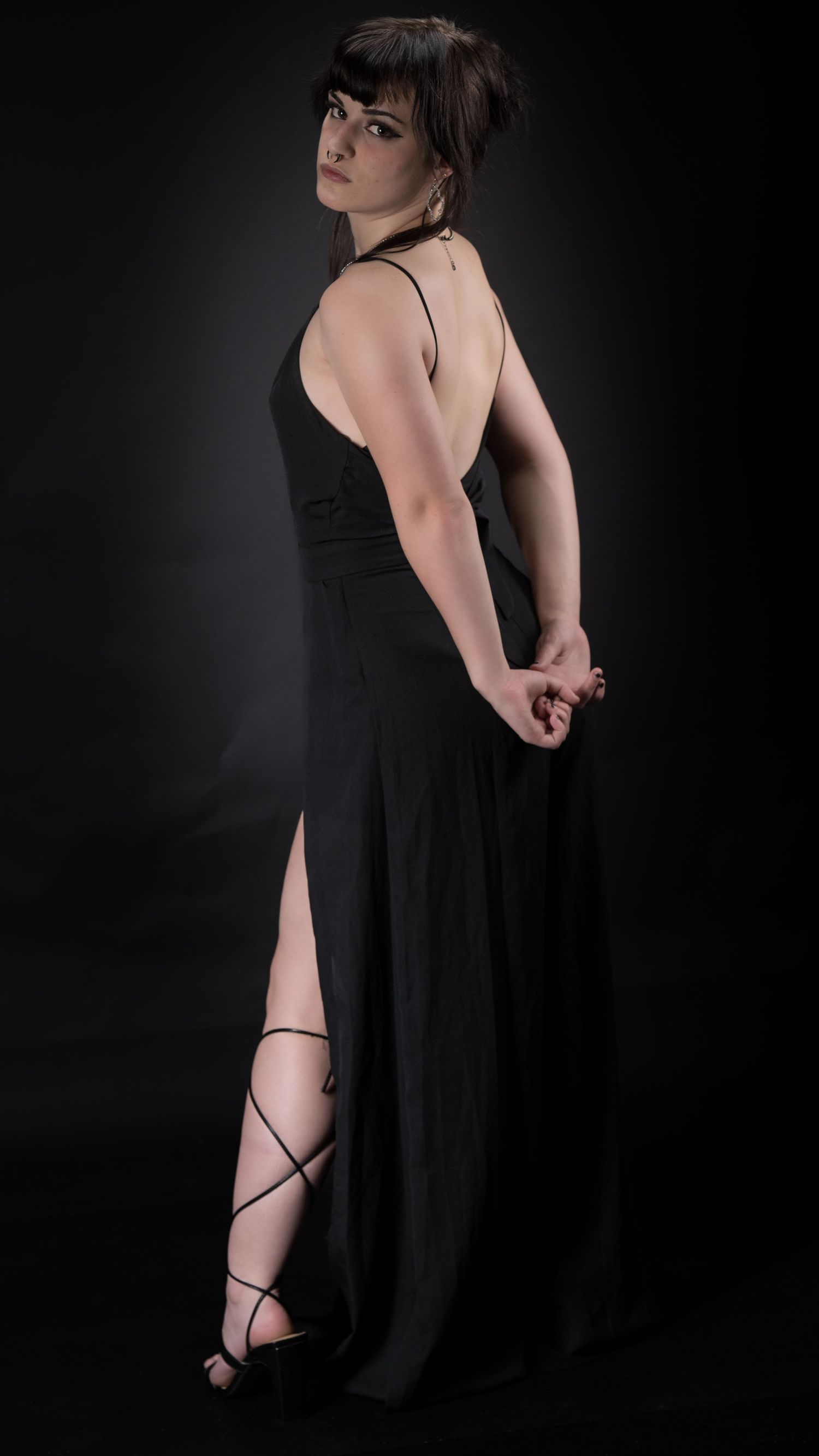Woman in elegant black dress and strappy sandals poses dramatically against dark background.