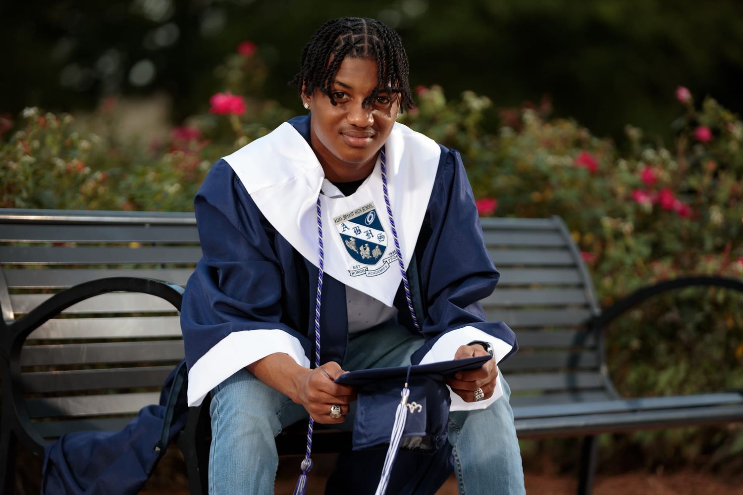 Student in navy varsity jacket reading while seated on park bench near rose garden.
