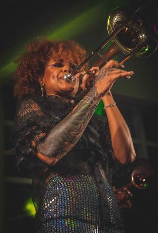 A musician playing trombone on stage in a sparkly outfit under dramatic lighting.