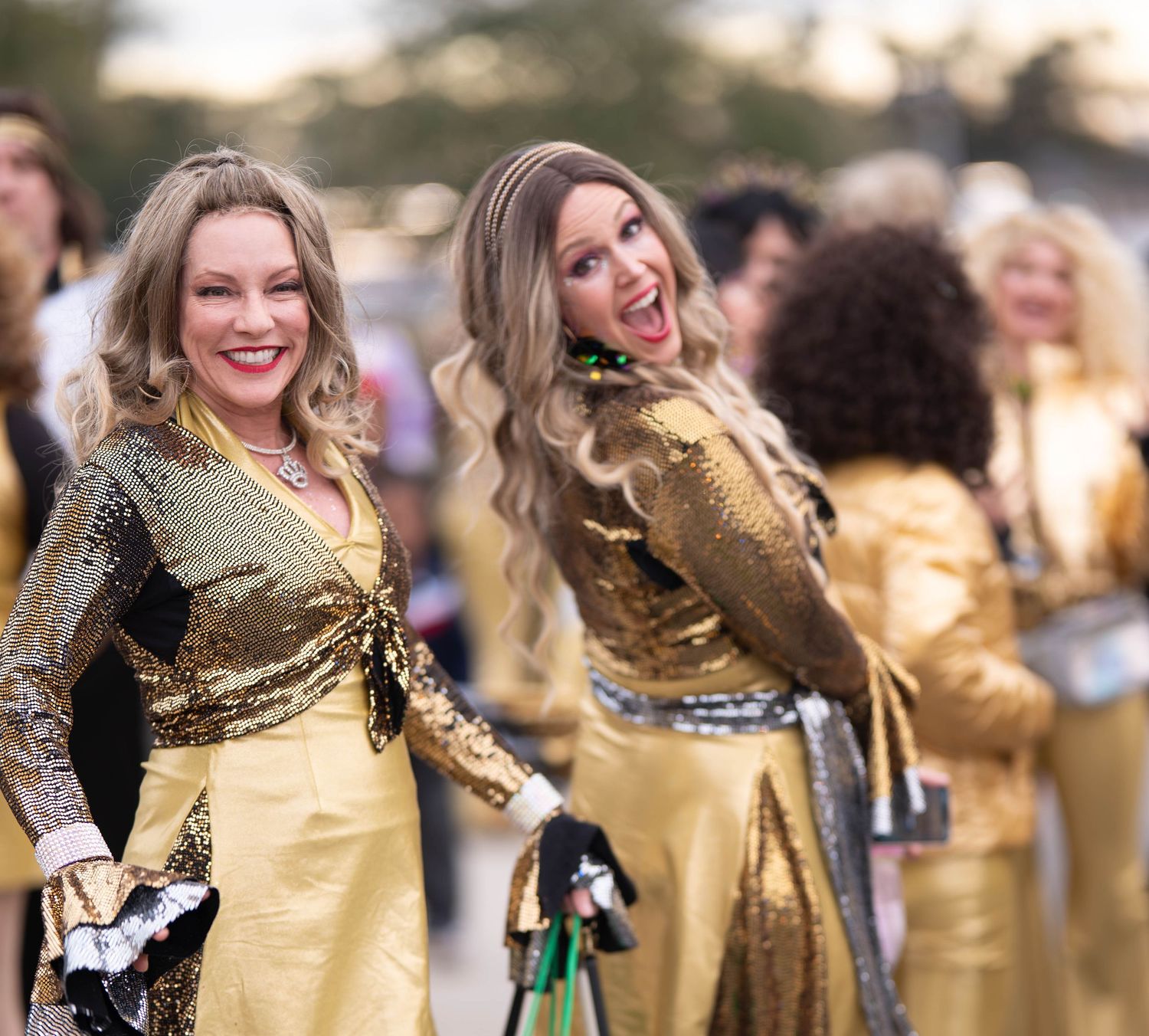 Two women in gold sequin outfits smiling at an outdoor event with others in matching costumes behind them.