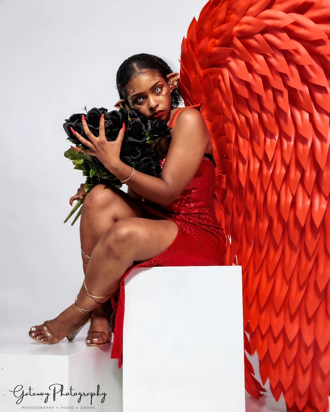 Artistic portrait with red wings holding flowers on white pedestal.