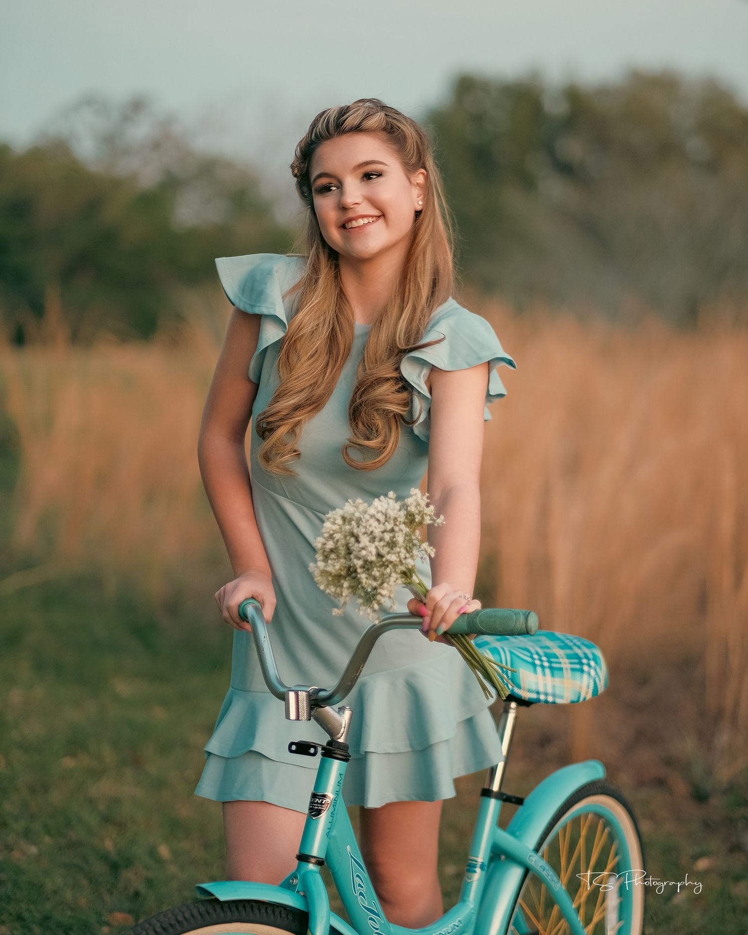 Smiling model in blue ruffled dress poses with vintage turquoise bicycle in rustic field setting.