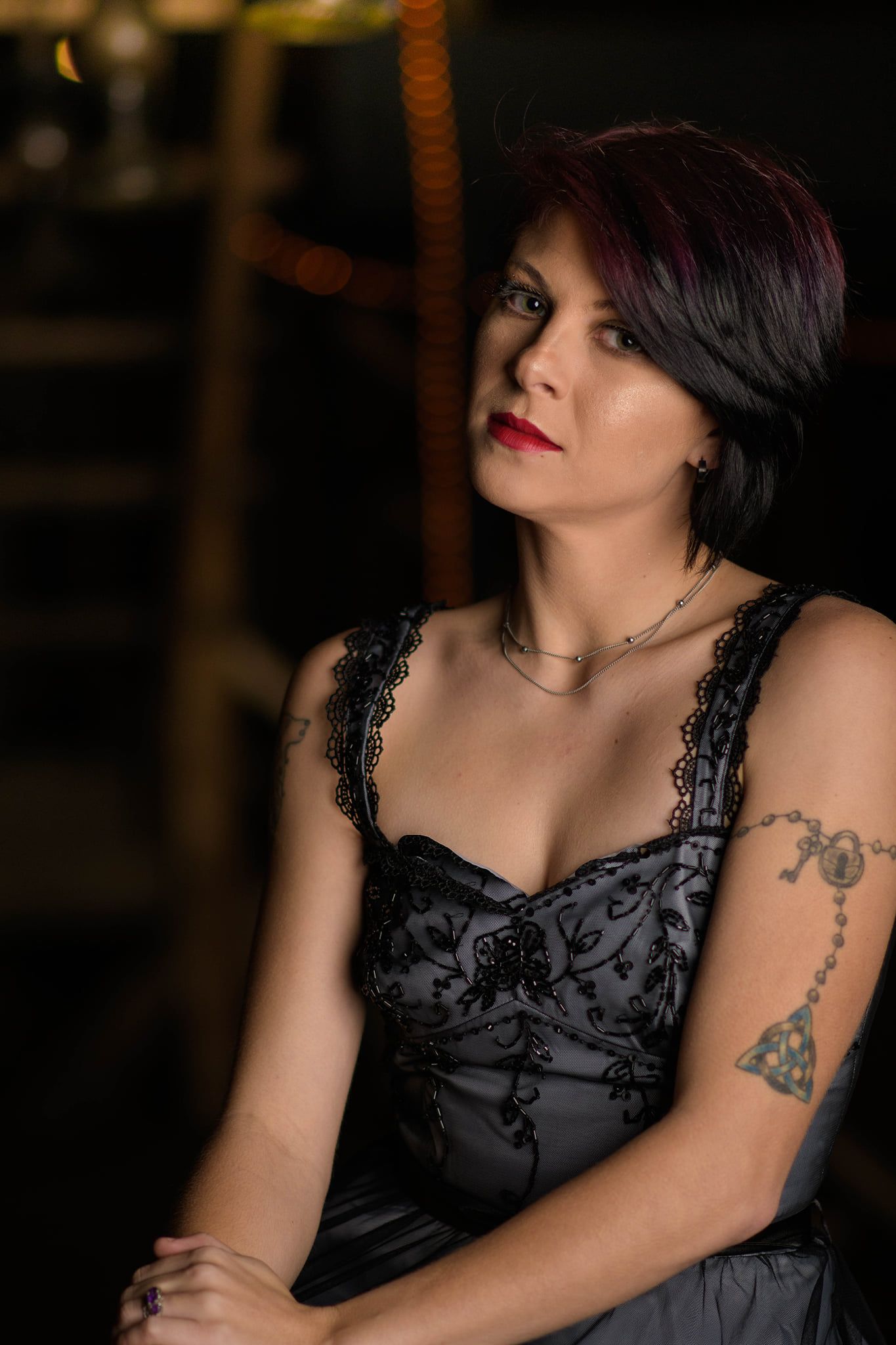 Portrait in a black lace dress with tattoos visible against moody lighting at night.