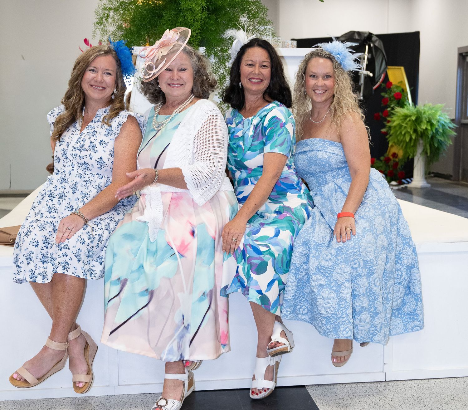 Four women in elegant floral dresses and fascinators pose together at a stylish garden party event.