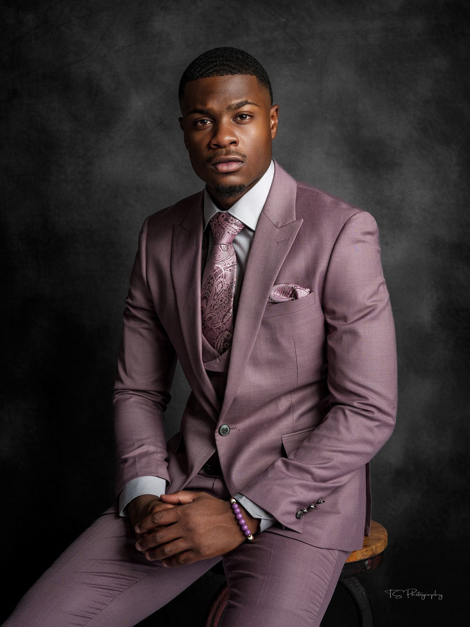 Professional headshot of person in mauve suit and patterned tie against grey backdrop.