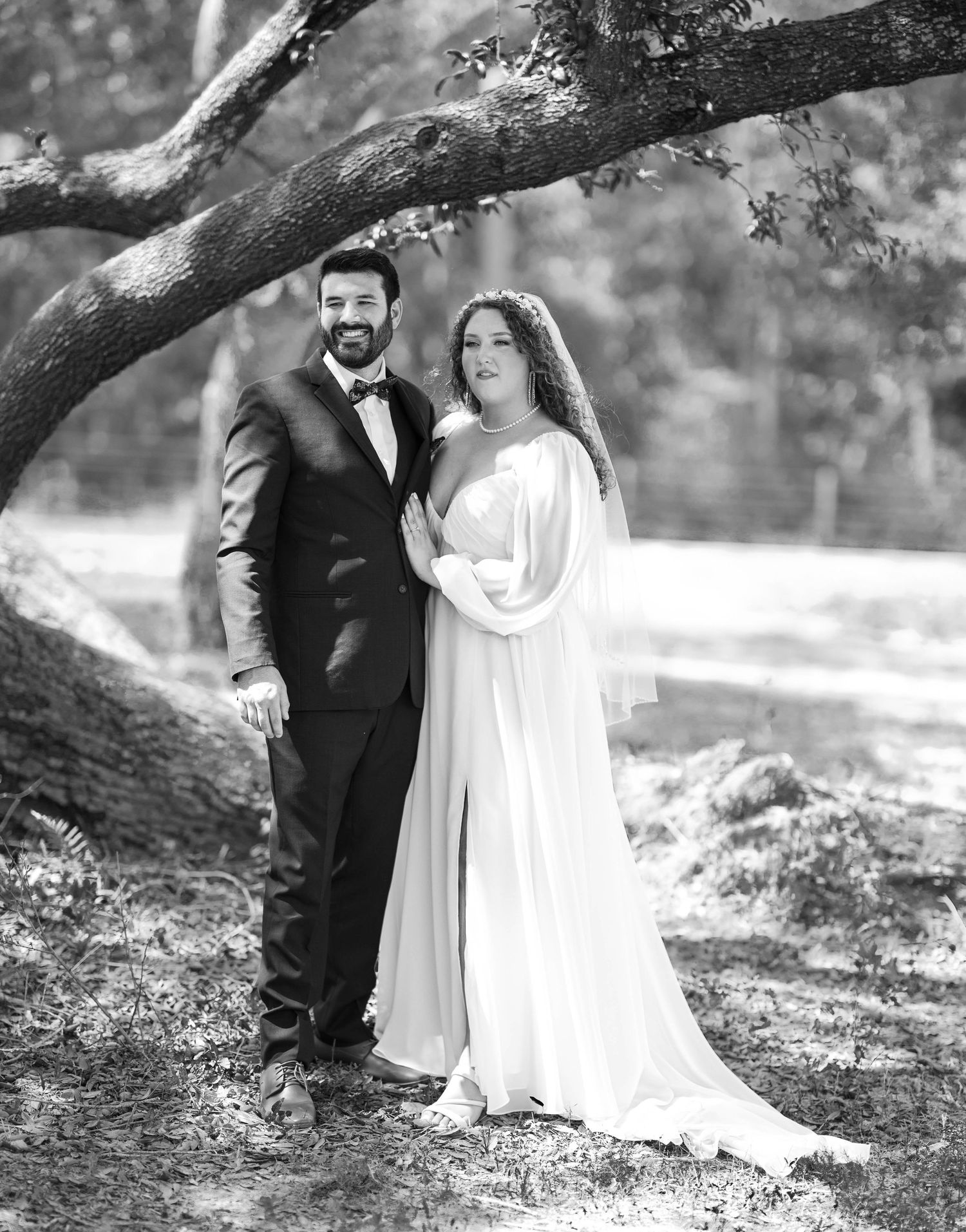A black and white wedding photograph shows a couple posing beneath a large tree branch in an outdoor setting.