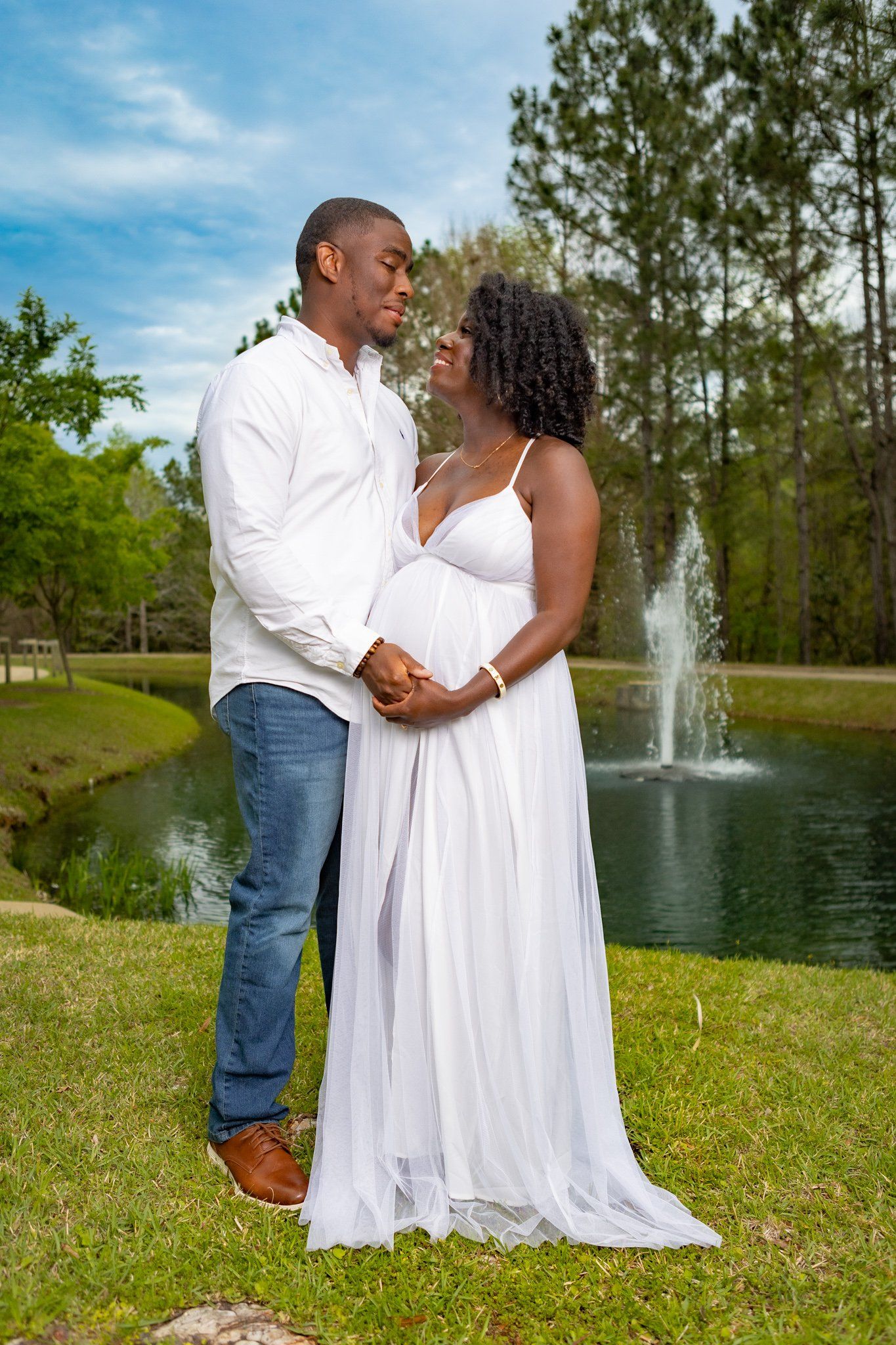 Romantic couple in white outfits posing by a fountain in a scenic park setting.