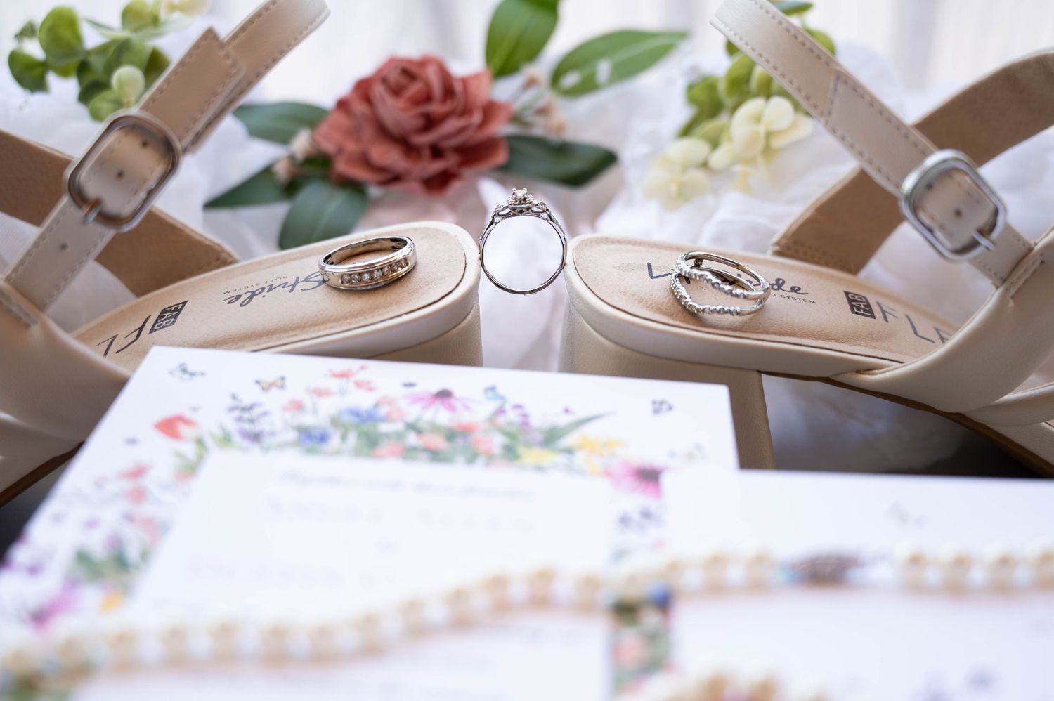 Wedding rings displayed between elegant beige heels with pink roses and floral stationery in the background.
