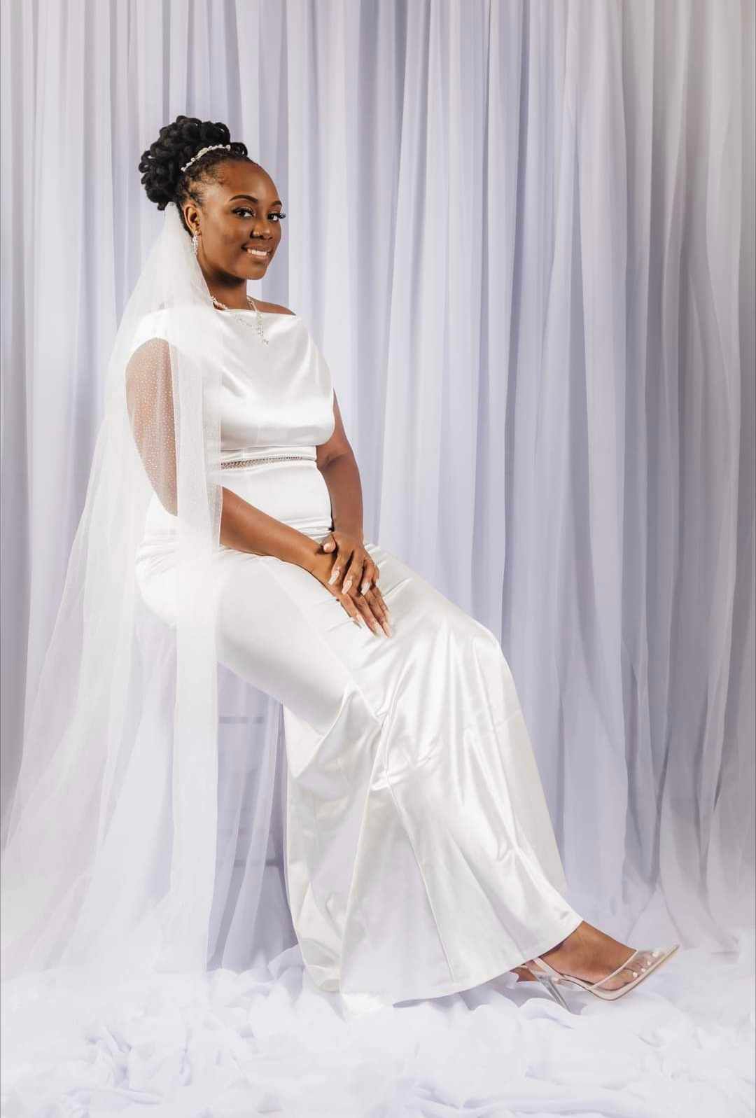 Elegant bridal portrait in modern white jumpsuit and veil against draped background.