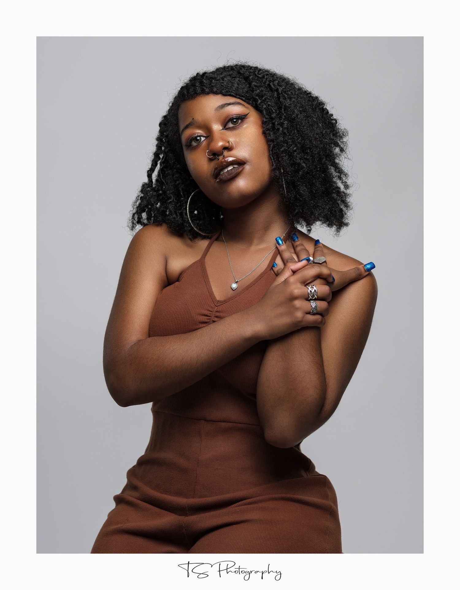 Studio portrait in brown dress with curly hair and jewelry against grey background.