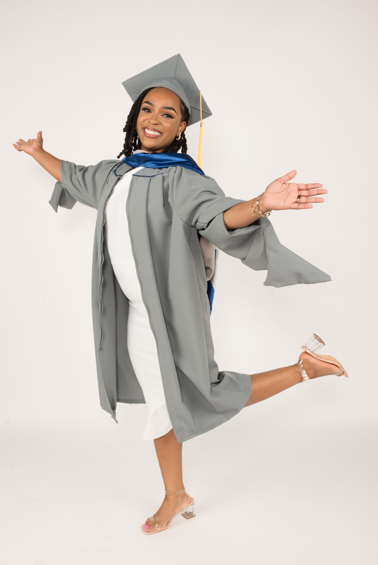 Joyful graduate in grey cap and gown strikes playful pose against white background.