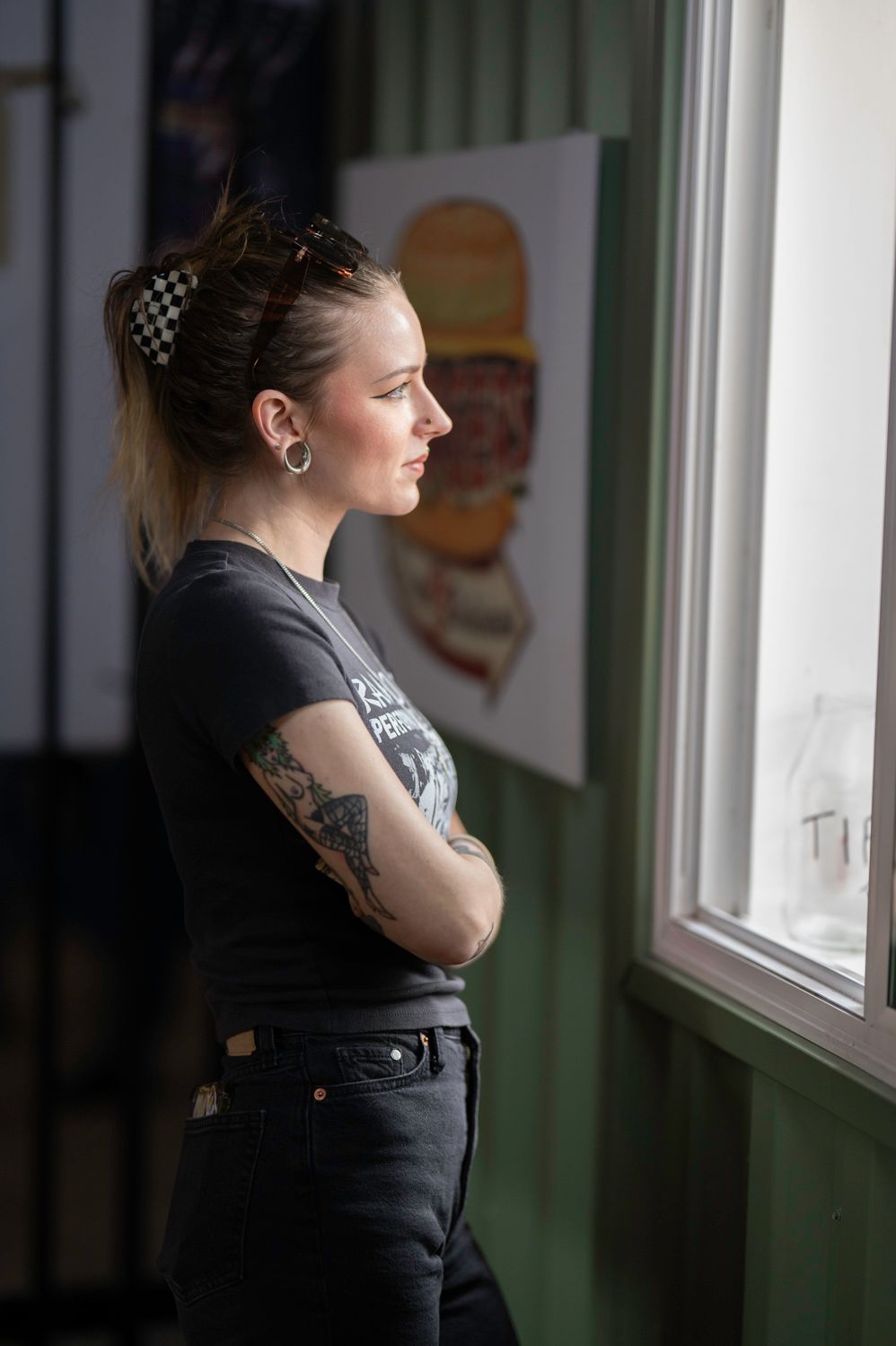 A person with tattoos and checkered hair accessories gazes through a window in casual black attire.