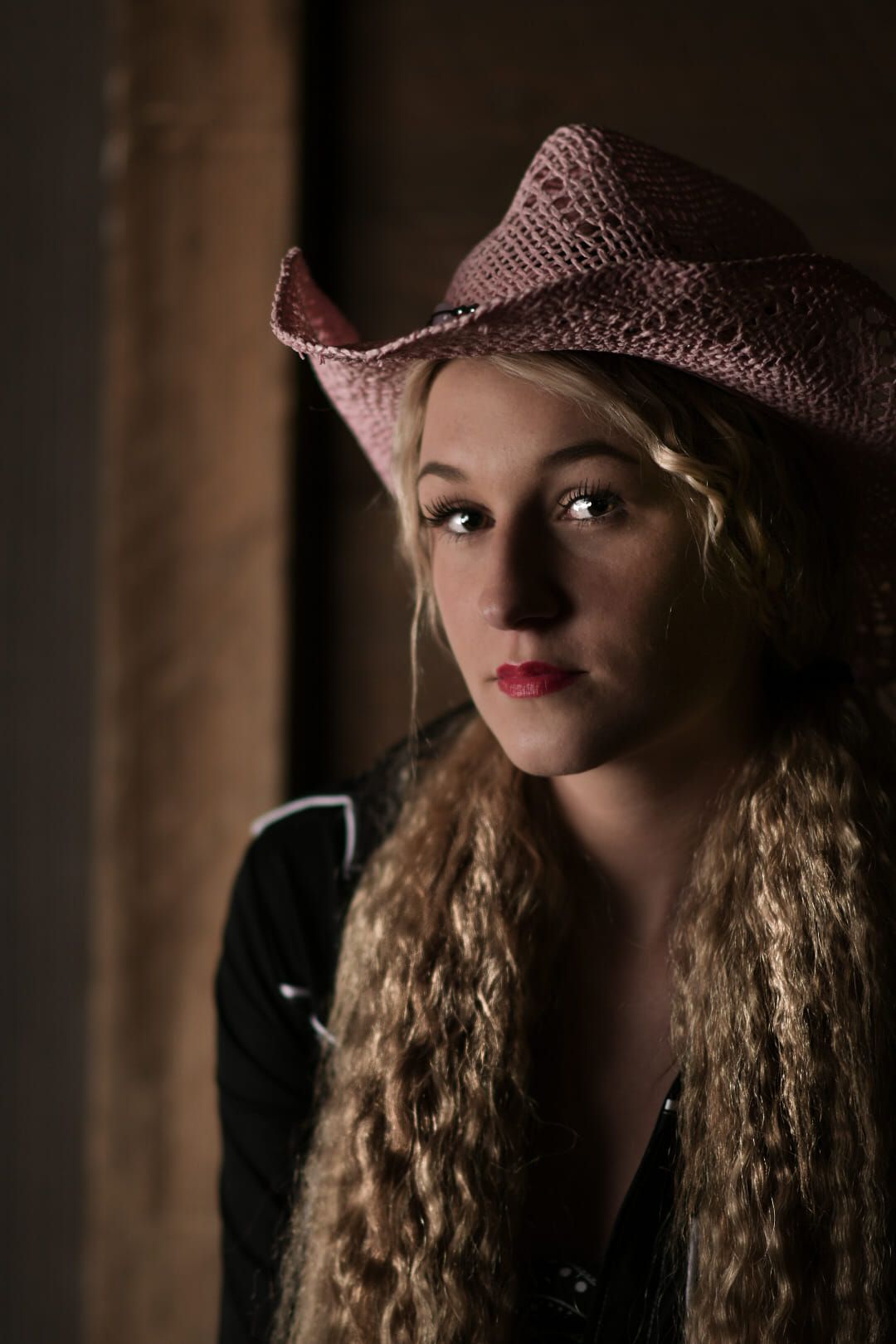 Portrait featuring long wavy hair and pink cowboy hat in dramatic low-key lighting with dark background.