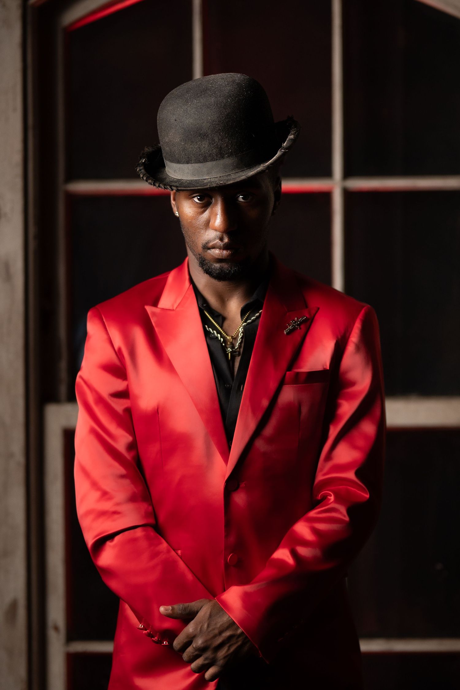 Dramatic portrait of person in red blazer and bowler hat against dark background.