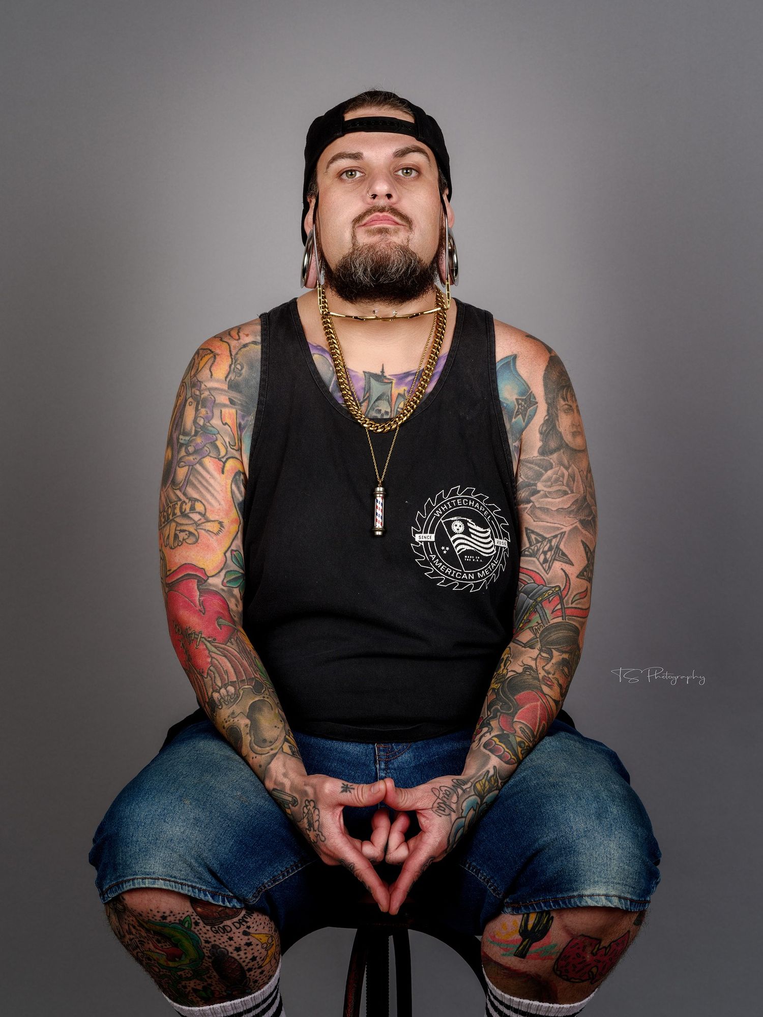 Tattooed model in black tank top and jeans seated on stool against grey background for portrait session.
