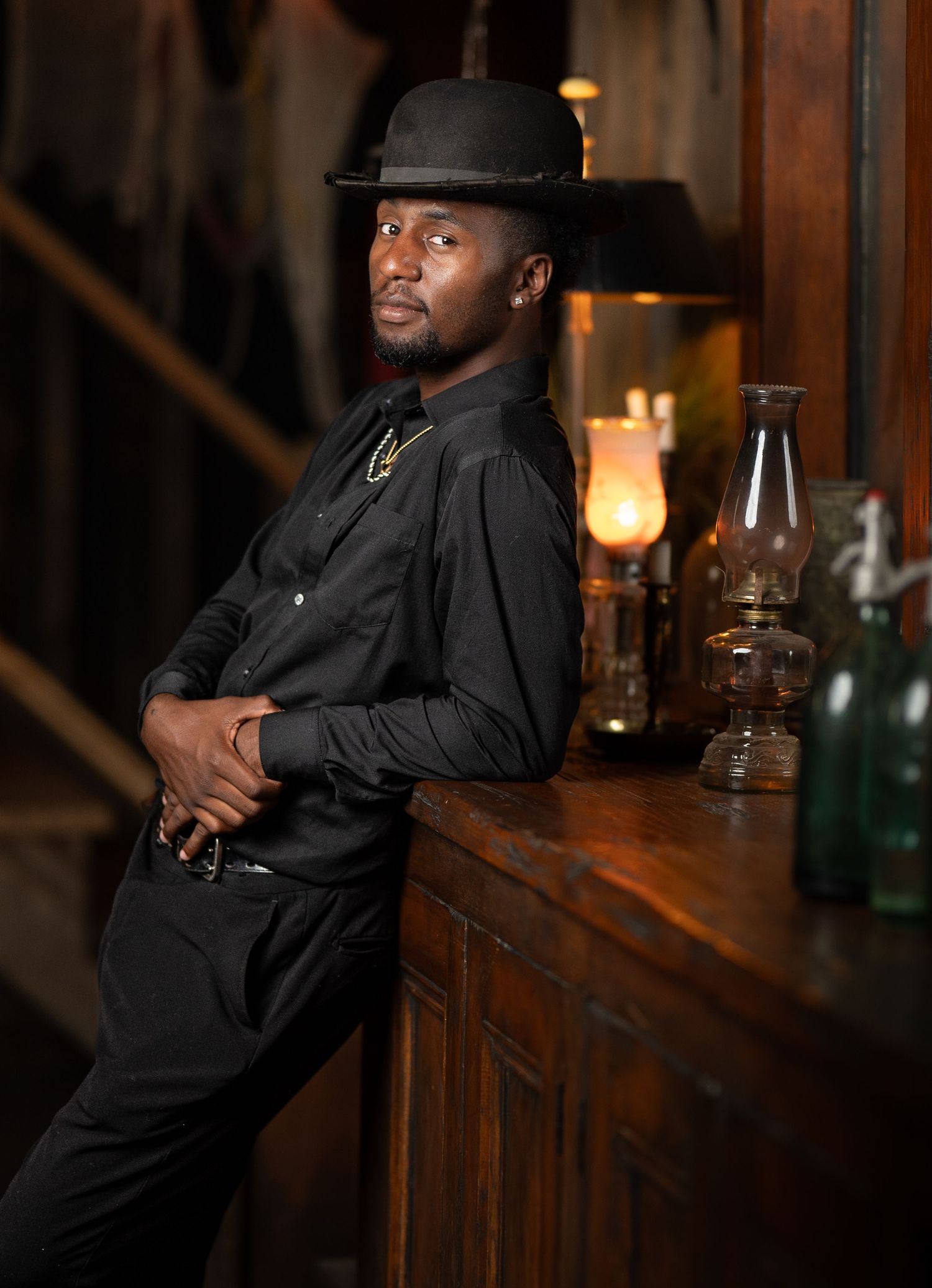 A person in black western attire and bowler hat leans against a rustic wooden bar with vintage glass lanterns and bottles.