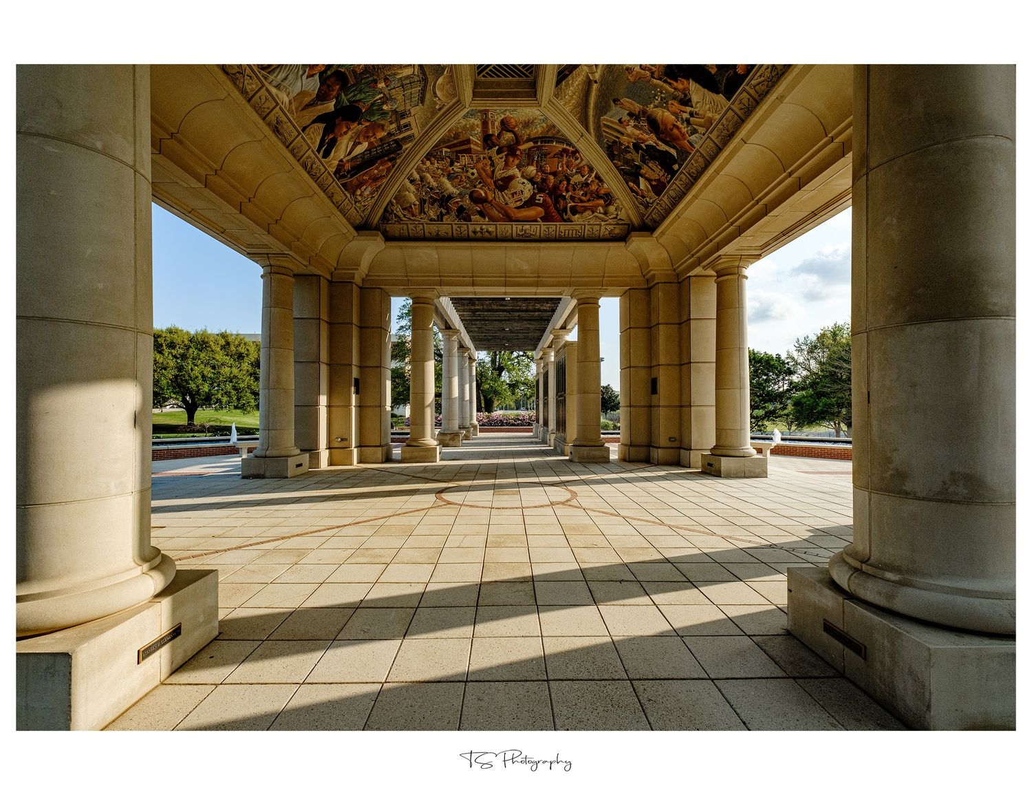 Architectural colonnade with ornate ceiling and stone pillars leading to a sun-lit outdoor space.