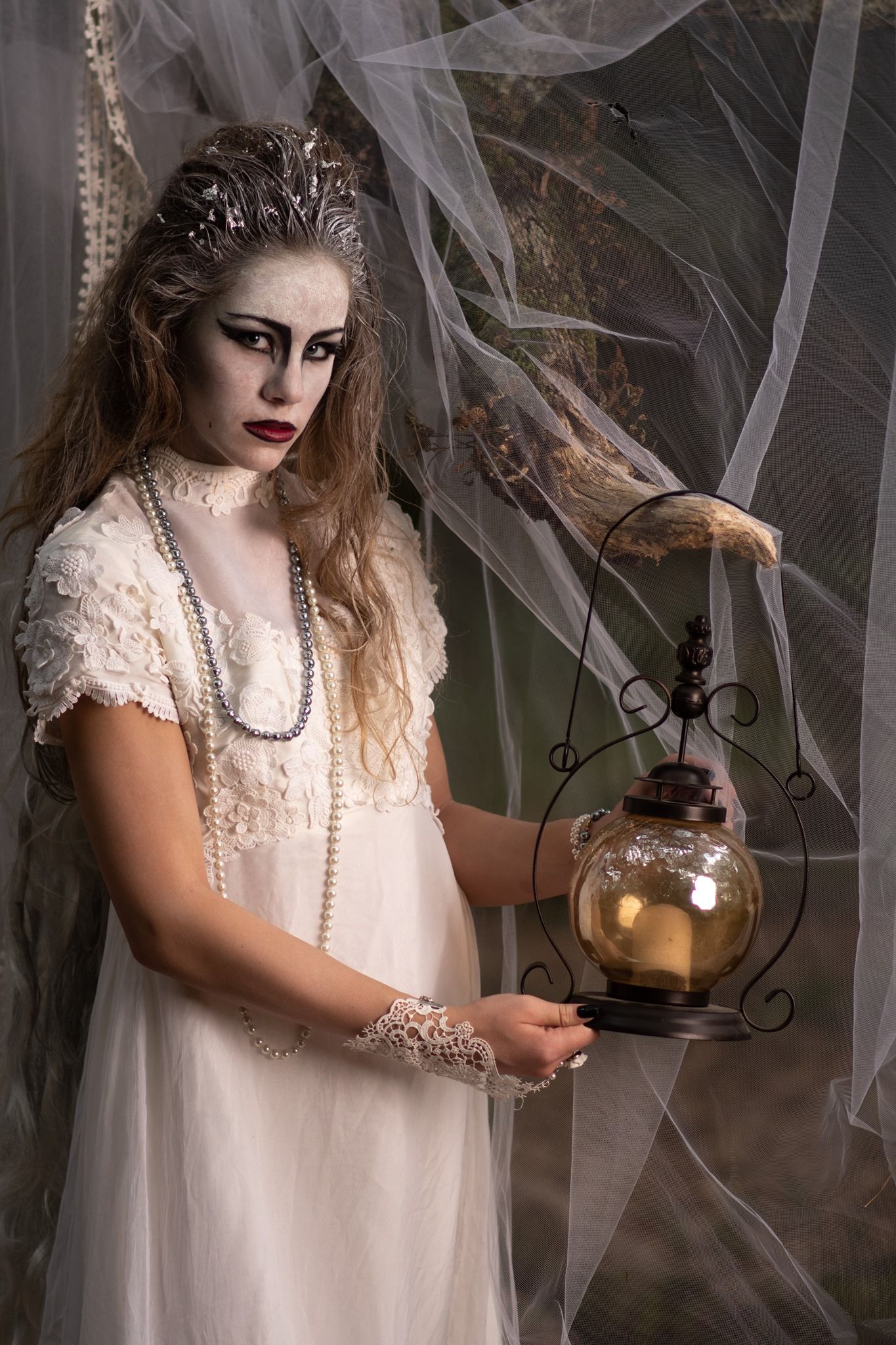 Gothic bride in vintage lace dress and pearls holds lantern among cobwebs in spooky themed photoshoot.
