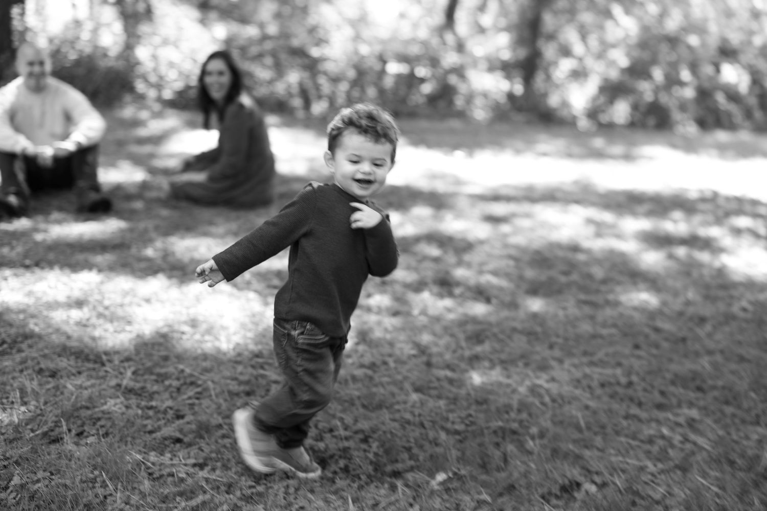 A child runs joyfully across a grassy park in black and white as adults sit in the background capturing motion.